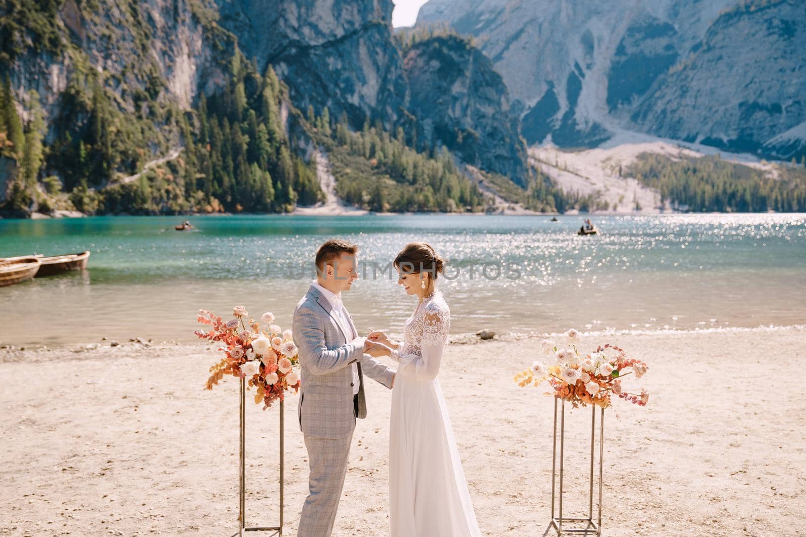 The bride dons a ring to the groom at the venue for the ceremony, with an arch of autumn floral columns, against the backdrop of Lago di Braies in Italy. Destination wedding in Europe, at Braies lake.