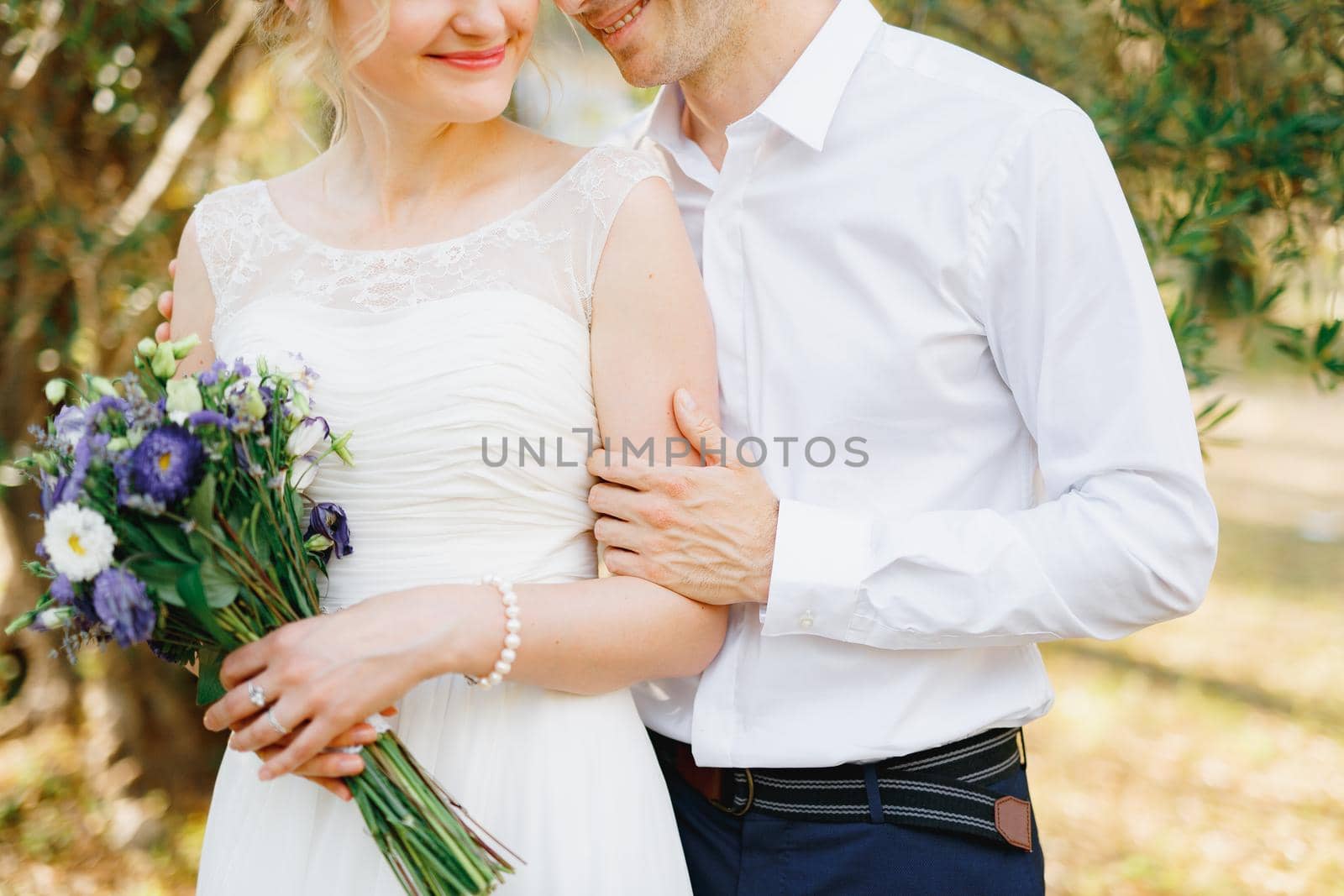 The groom gently hugs the bride near the green trees in the olive grove, the bride holds a bouquet of blue flowers by Nadtochiy