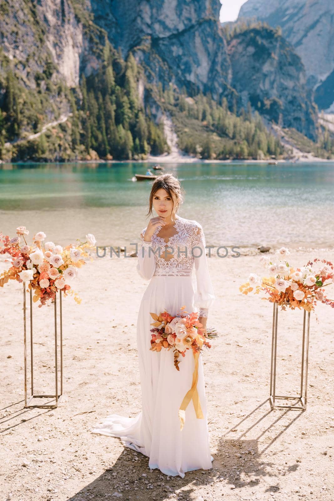 Beautiful bride in a white dress with sleeves and lace, with a yellow autumn bouquet on background of the arch for ceremony, at Lago di Braies in Italy. Destination wedding in Europe, on Braies lake. by Nadtochiy