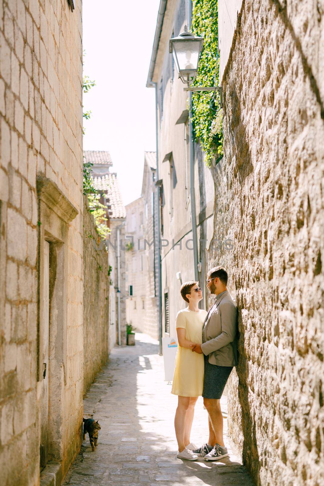 A man and a woman hugging at a white brick wall in a narrow alley of the old town of Perast by Nadtochiy