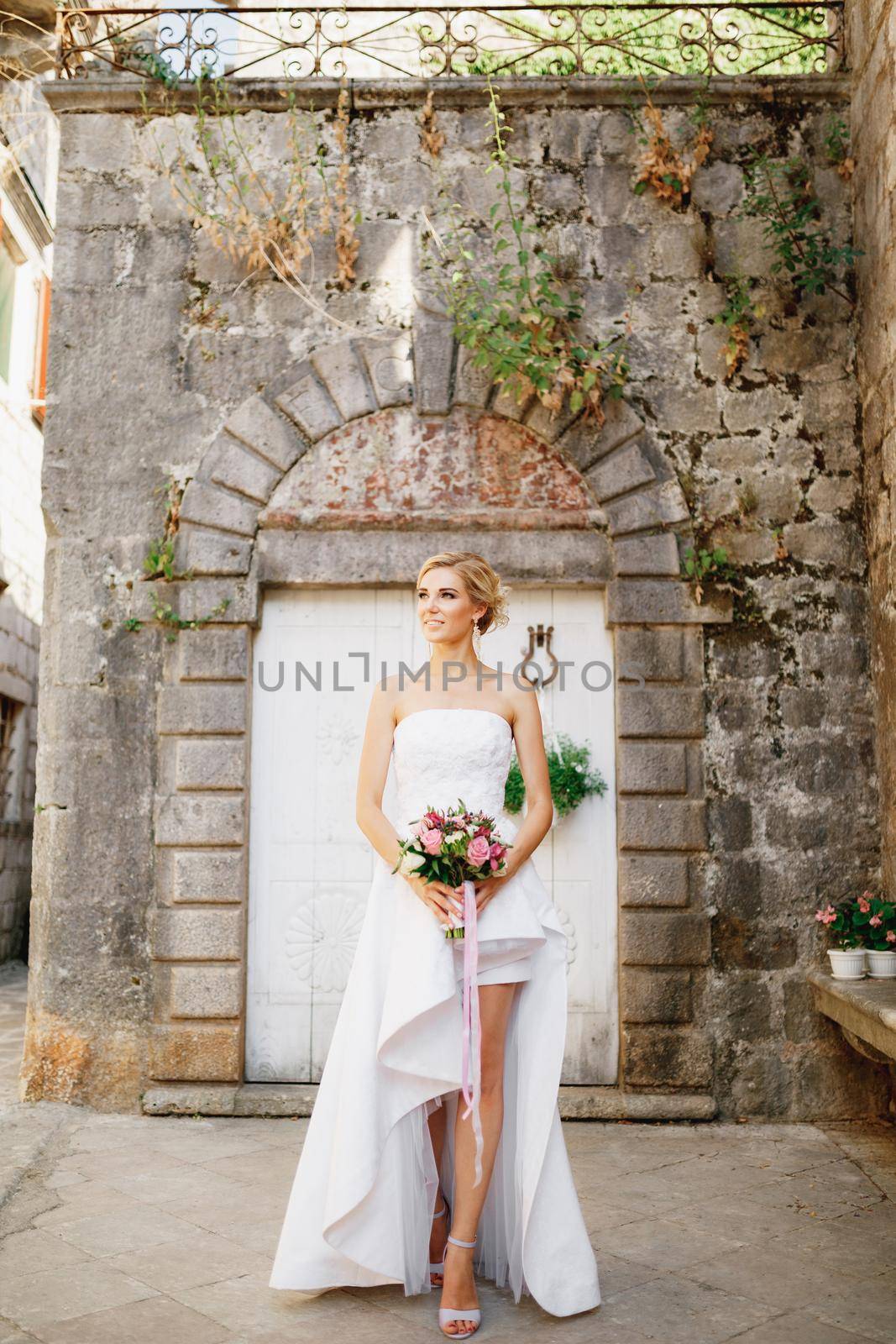 A bride in an elegant dress with a bouquet in her hands stands at the white doors of a beautiful old building in Perast by Nadtochiy