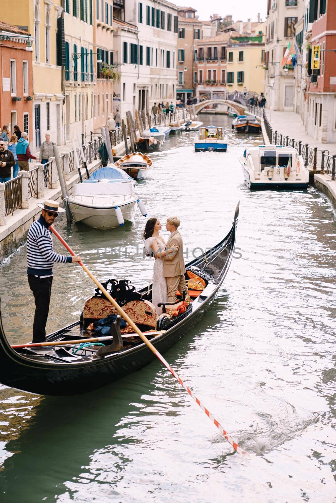 Venice, Italy - 04 october 2019: Italy wedding in Venice. A gondolier rolls a bride and groom in a classic wooden gondola along a narrow Venetian canal. Newlyweds are standing in the boat, rear view. by Nadtochiy