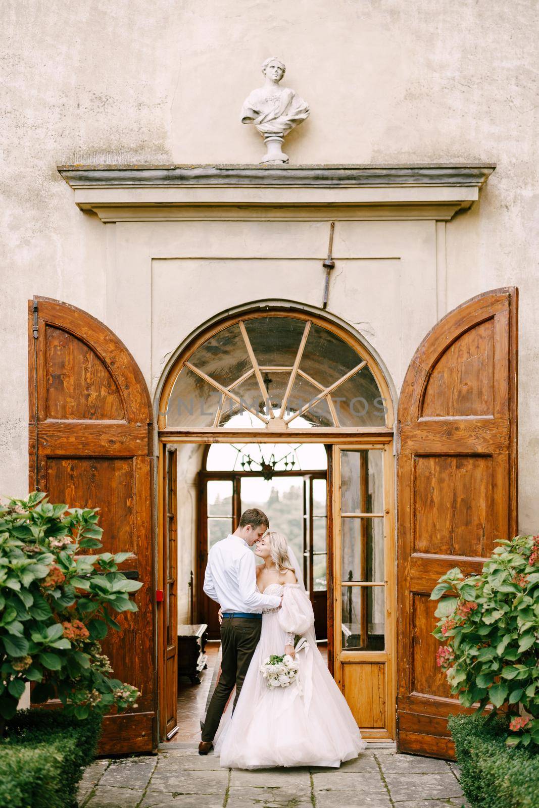 Wedding at an old winery villa in Tuscany, Italy. A wedding couple stands near the old wooden doors at the villa-winery.