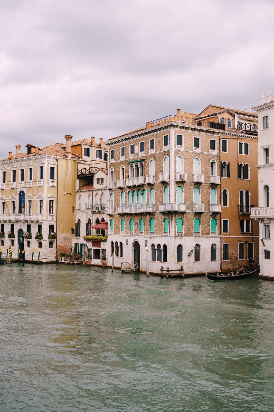A gondola with tourists is sailing along Grand Canal amid the facades of Venetian houses standing on the water in Venice, Italy.