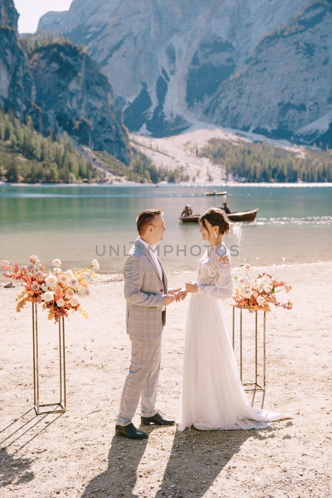 The groom dons a bride's ring, in place for the ceremony, with an arch of autumn floral columns, against the backdrop of Lago di Braies in Italy. Destination wedding in Europe, at Braies lake.