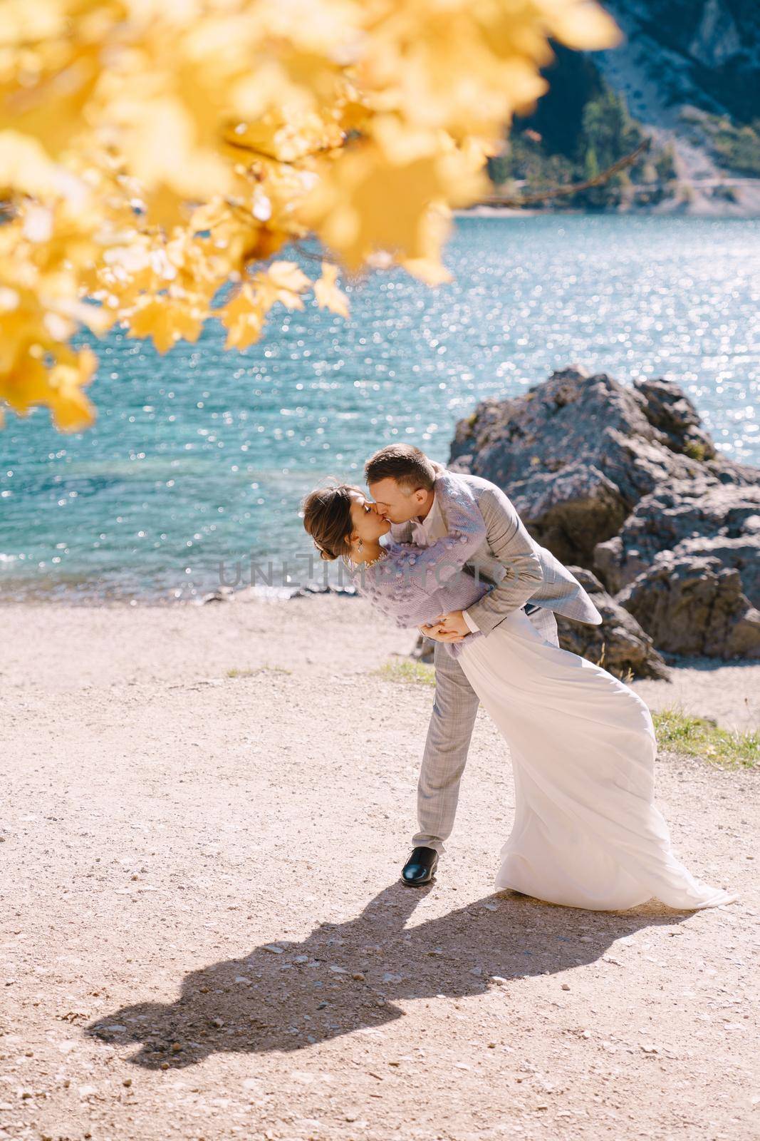 Bride and groom under an autumn tree, with fiery yellow foliage, at the Lago di Braies in Italy. Destination wedding in Europe, on Braies lake. Lovers newlyweds kiss in a dance. by Nadtochiy