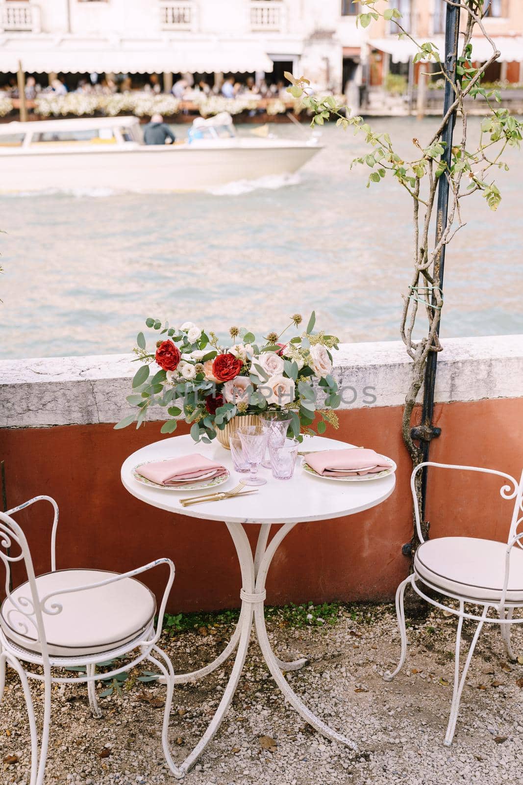 A white table and chairs for two, in a restaurant on the banks of the Grand Canal in Venice, Italy. On the table there is large bouquet of roses and leaves of eucalyptus, glasses, plates and cutlery
