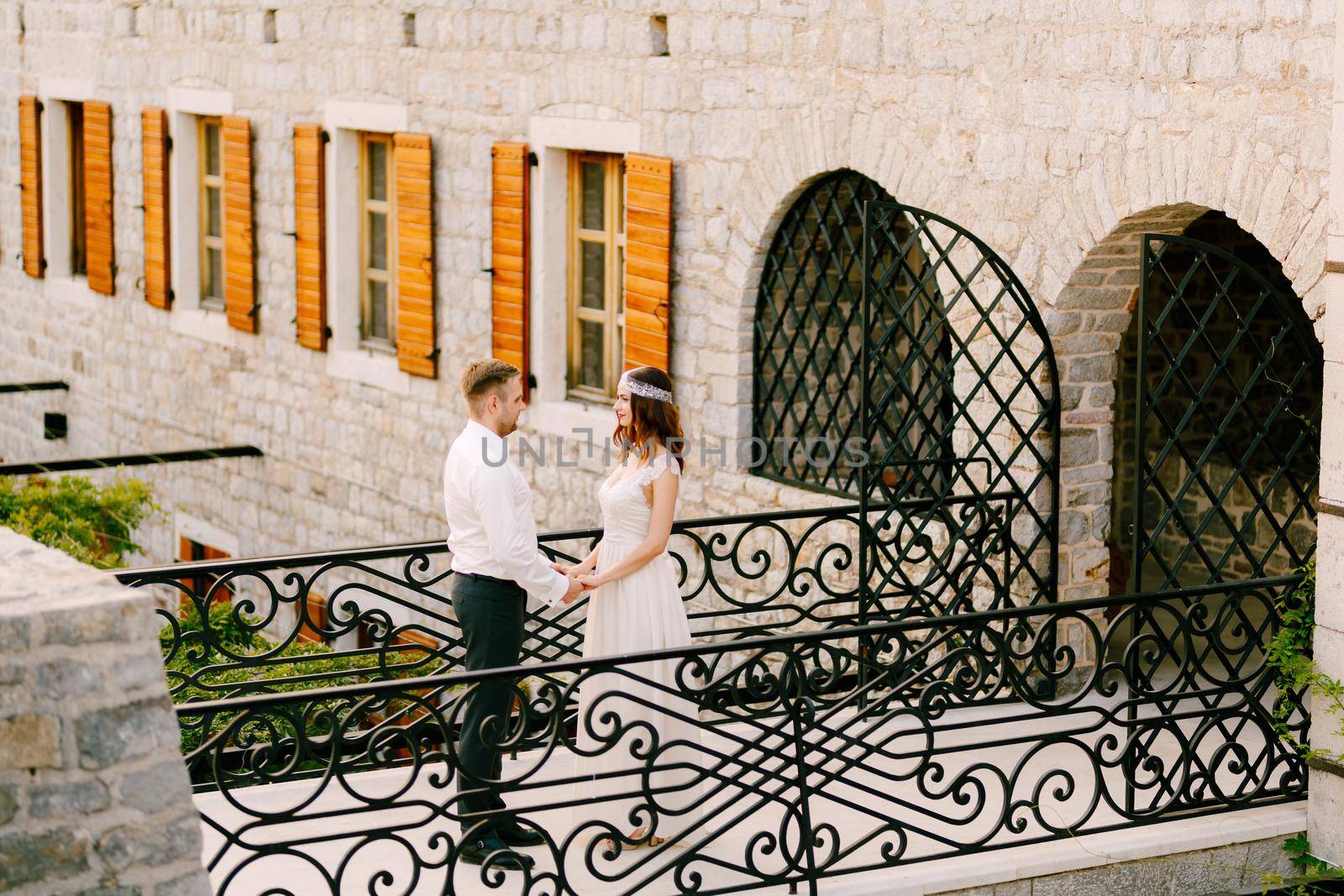 The bride and groom stand on a beautiful forged bridge in the old town of Budva holding hands and looking into each other's eyes . High quality photo