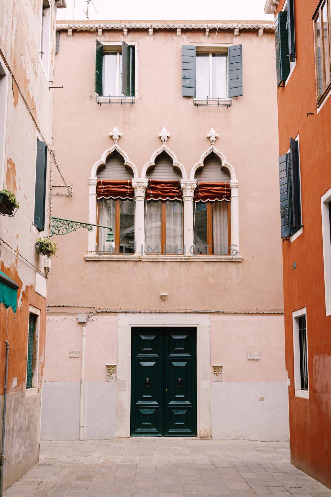 The facade of a three-story peach house in Venice, Italy. Massive green wooden front door, three classic Venetian windows with a sharp top, columns and rag rolls.