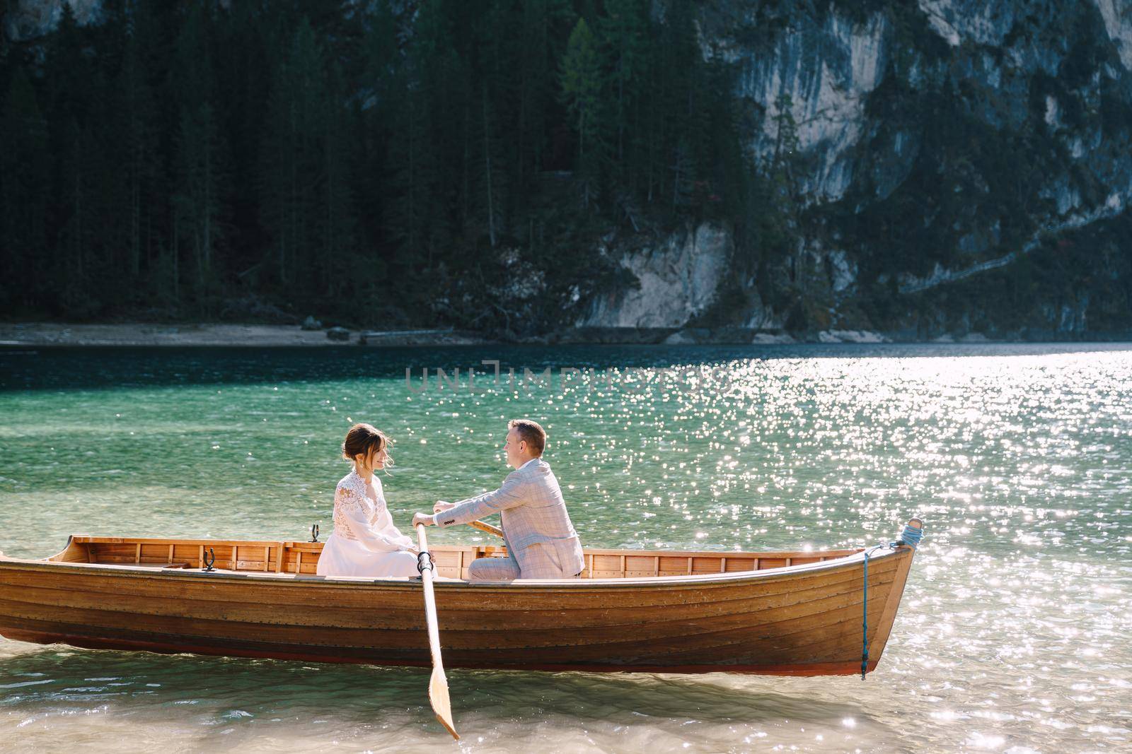 Newlyweds sail in a wooden boat on the Lago di Braies in Italy. Wedding in Europe, on Braies lake. Wedding couple - The groom rows with wooden oars, the bride sits opposite. by Nadtochiy