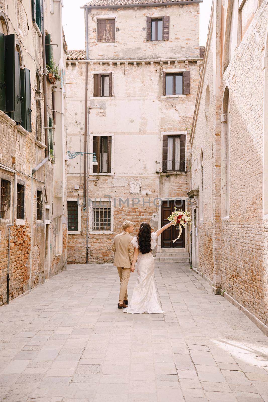 Italy wedding in Venice. The bride and groom walk along the deserted streets of the city. Newlyweds are walking in a dead end alley on the background of brick buildings. by Nadtochiy