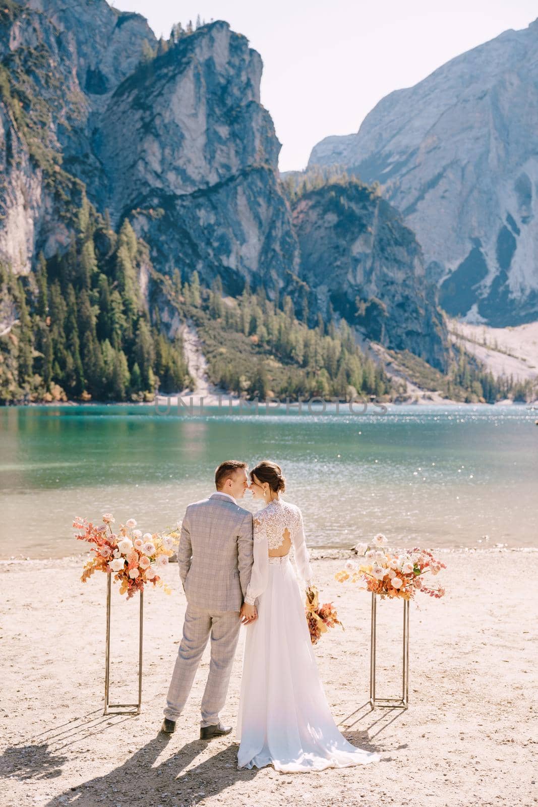 The bride and groom stand still for the ceremony, with an arch of autumn floral columns, against the backdrop of Lago di Braies in Italy. Destination wedding in Europe, at Braies lake.