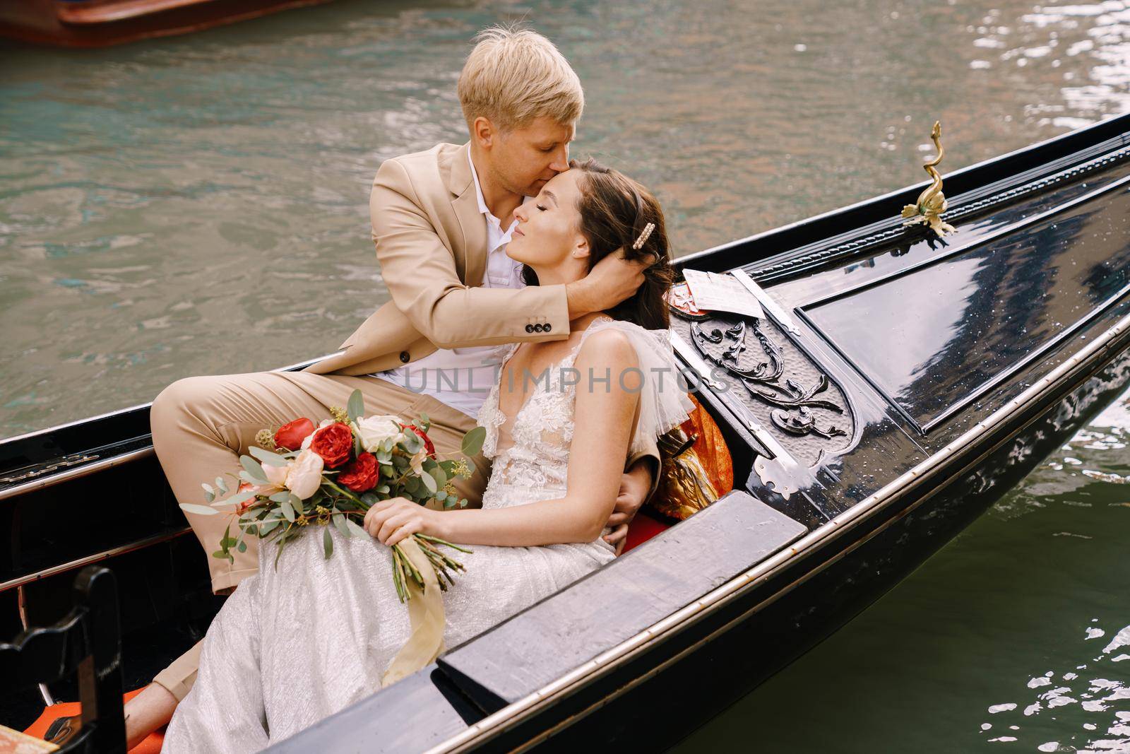 The bride and groom ride in a classic wooden gondola along a narrow Venetian canal. A close-up of cuddling newlyweds.