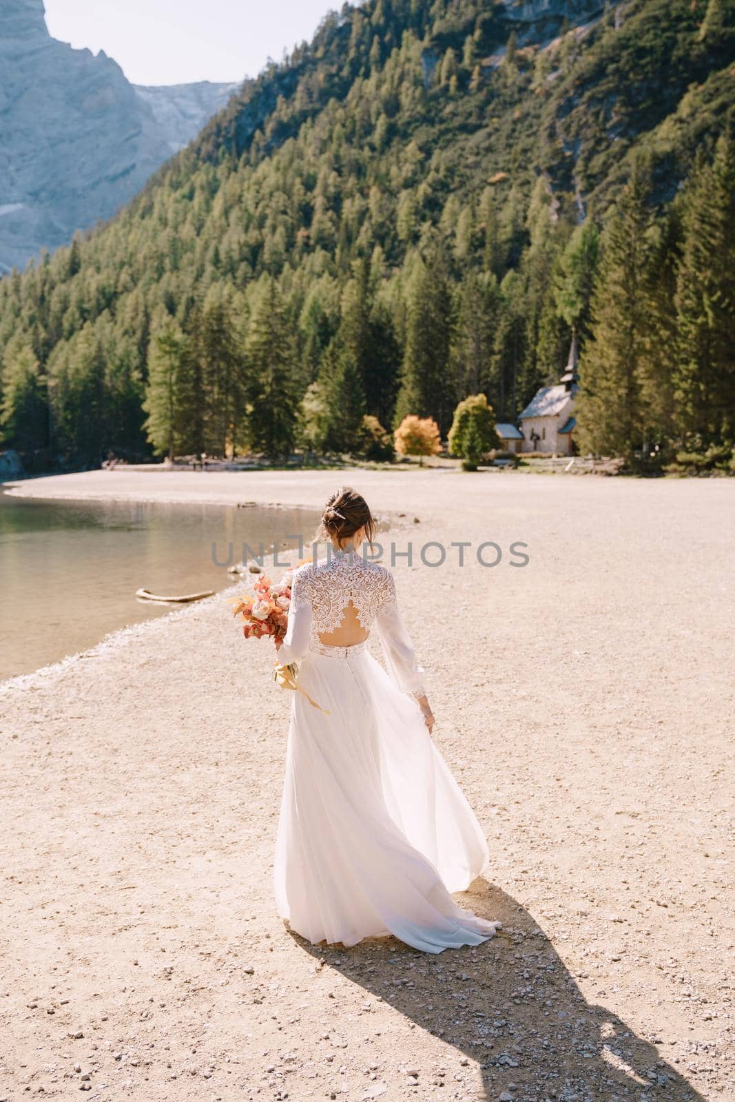 Beautiful bride in a white dress with sleeves and lace, with a yellow autumn bouquet of dried flowers and peony roses, at Lago di Braies in Italy. Destination wedding in Europe, at Braies lake.