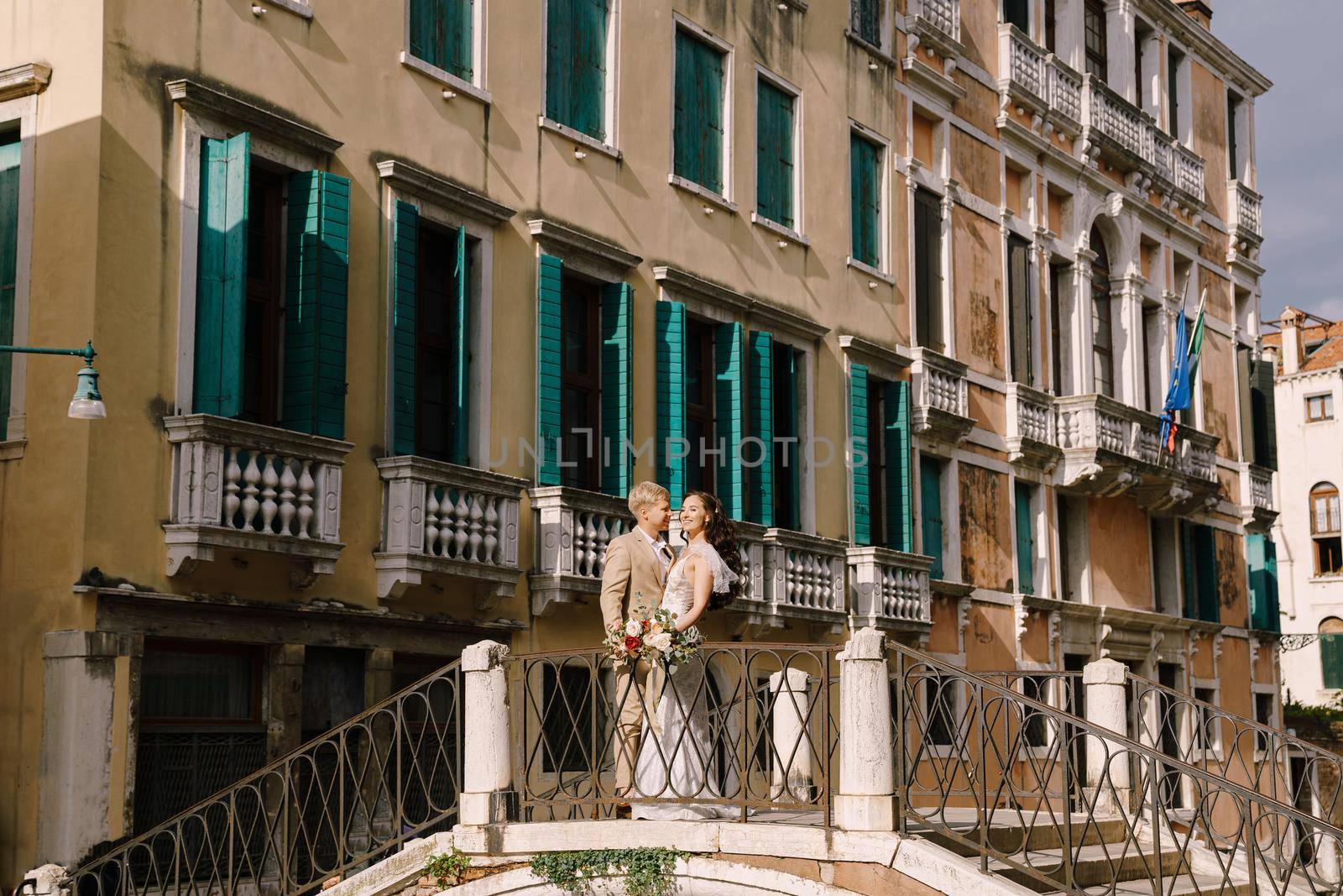 Italy wedding in Venice. The bride and groom are standing on a stone bridge over a narrow Venetian canal. Newlyweds walk around the city taking pictures on the street. by Nadtochiy