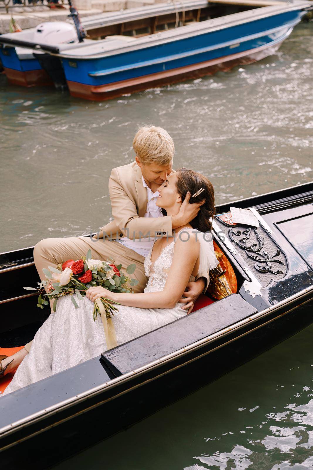 The bride and groom ride in a classic wooden gondola along a narrow Venetian canal. A close-up of cuddling newlyweds.