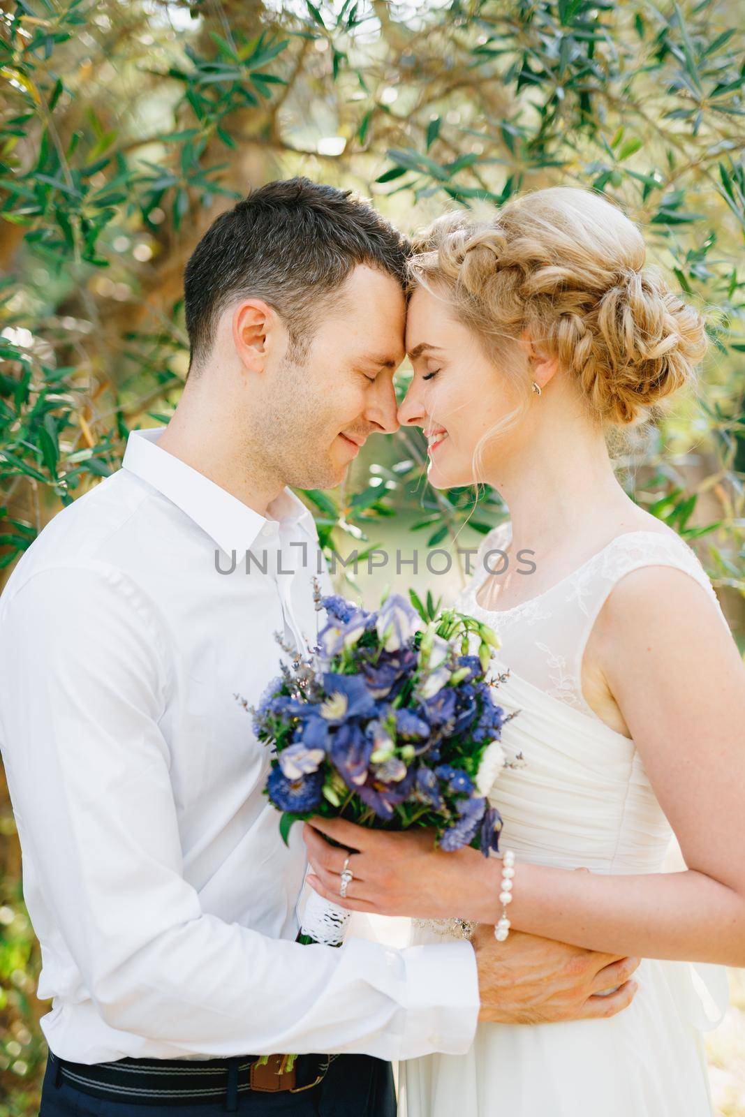 Bride with a bouquet of blue flowers and the groom embracing tenderly in the olive grove by Nadtochiy