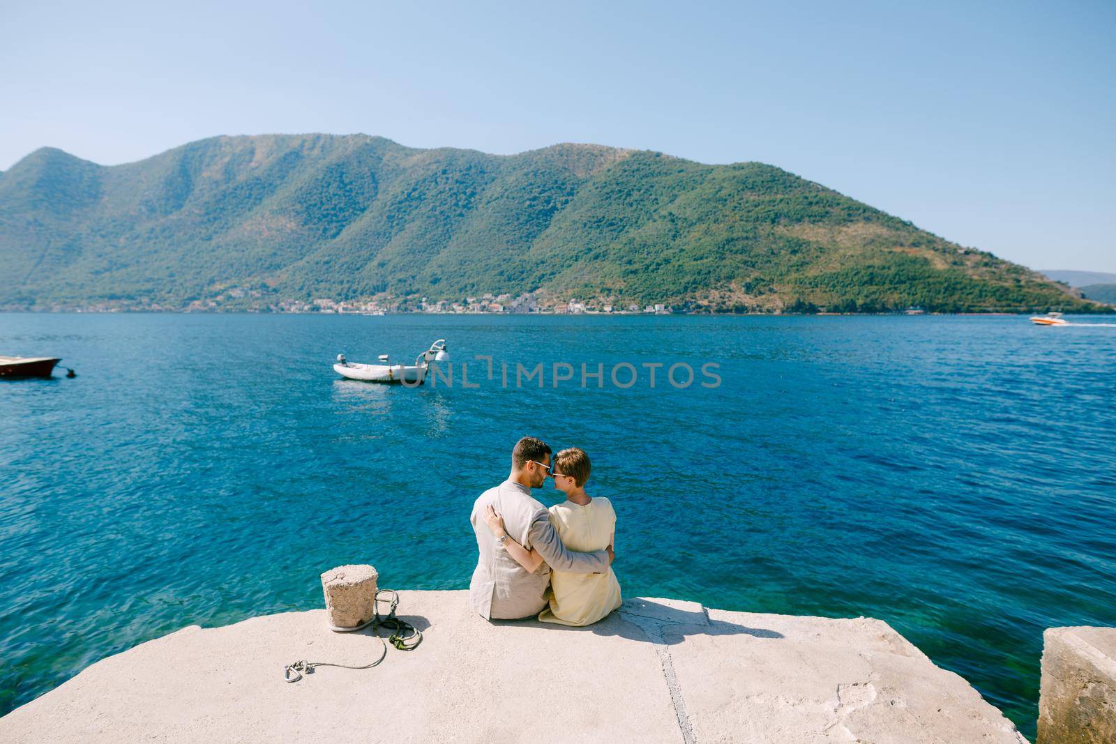 A man and a woman sitting and hugging on the edge of a pier near Perast in the Bay of Kotor, back view by Nadtochiy