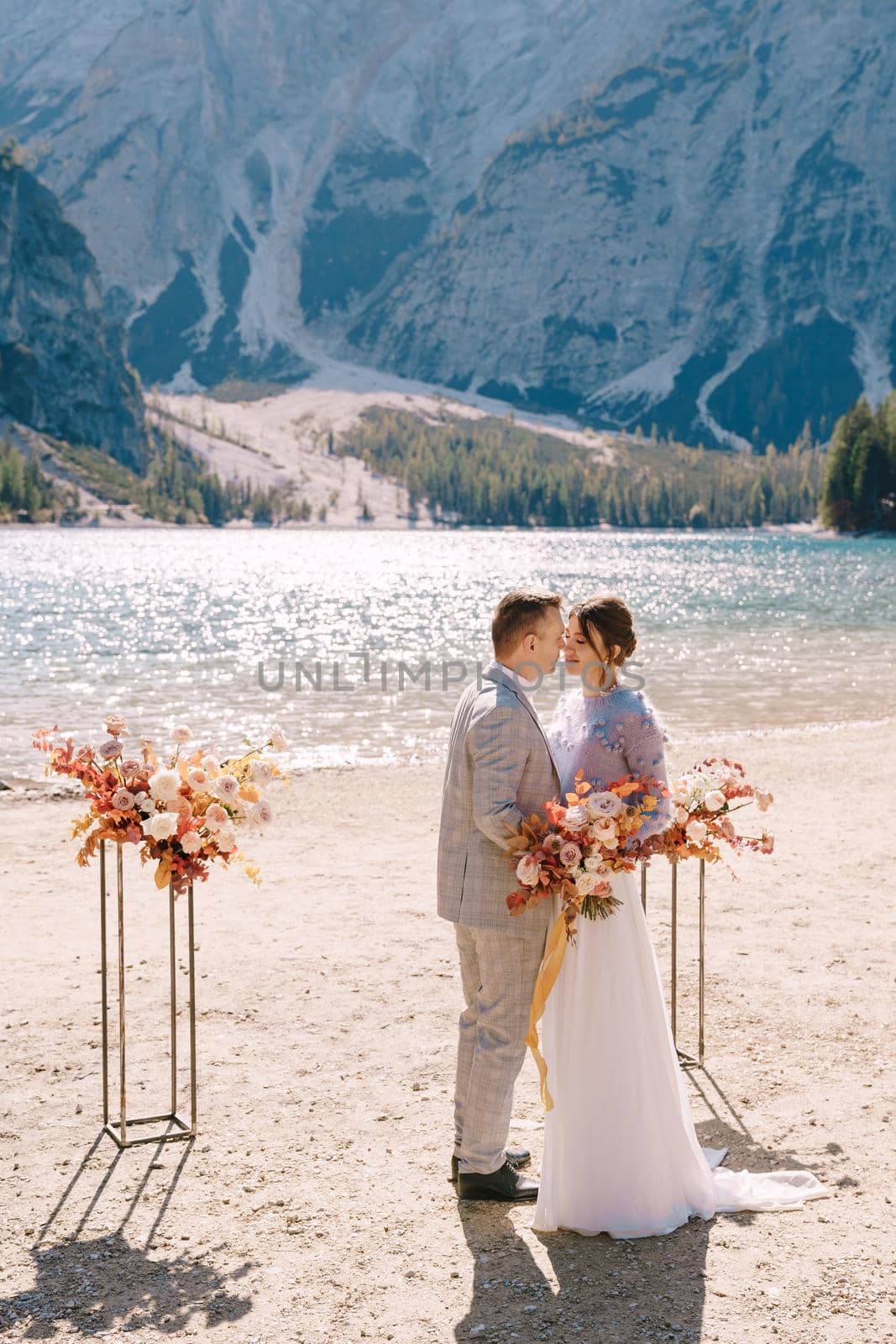 The groom dons a bride's ring, in place for the ceremony, with an arch of autumn floral columns, against the backdrop of Lago di Braies in Italy. Destination wedding in Europe, at Braies lake.