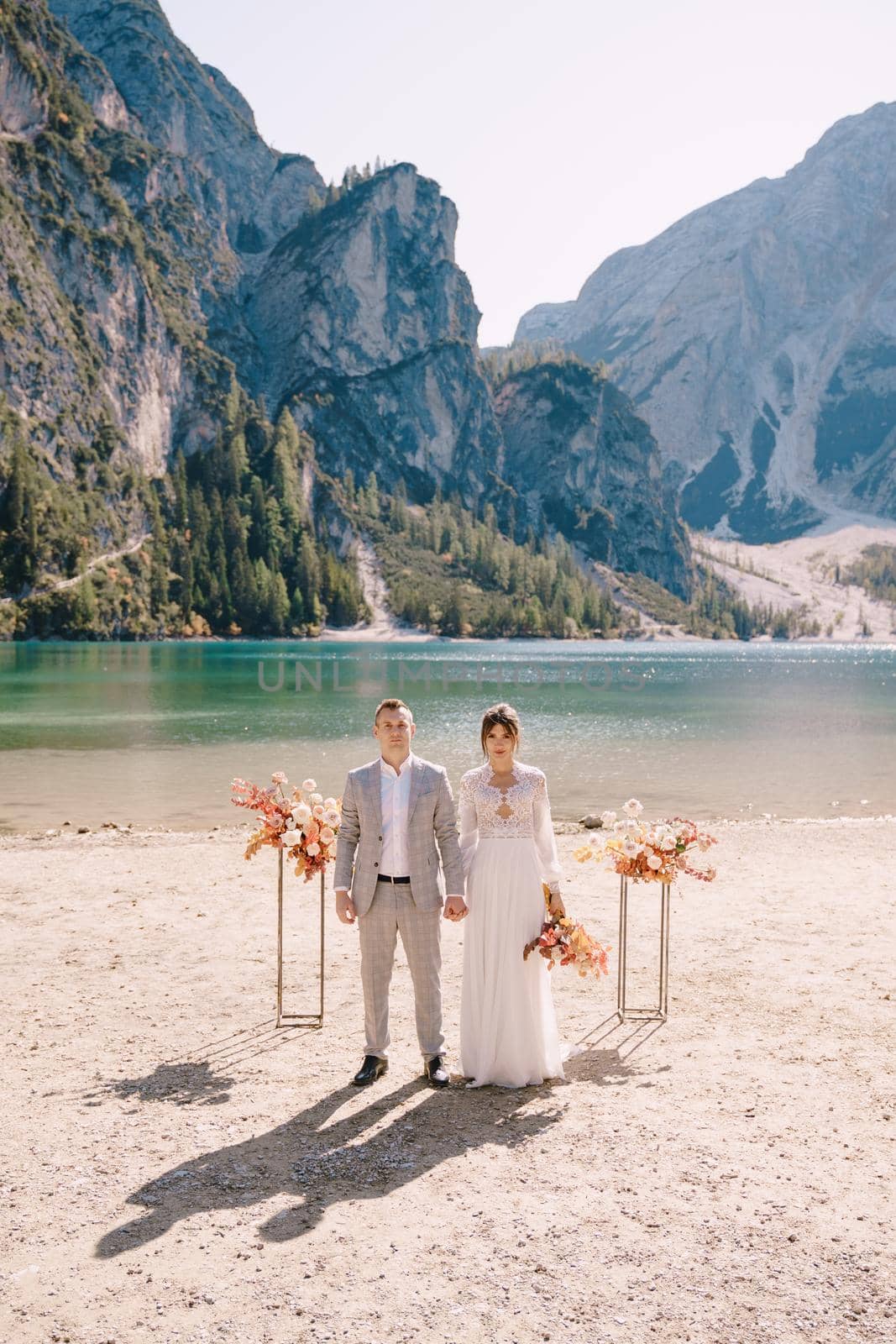 The bride and groom stand still for the ceremony, with an arch of autumn floral columns, against the backdrop of Lago di Braies in Italy. Destination wedding in Europe, at Braies lake.