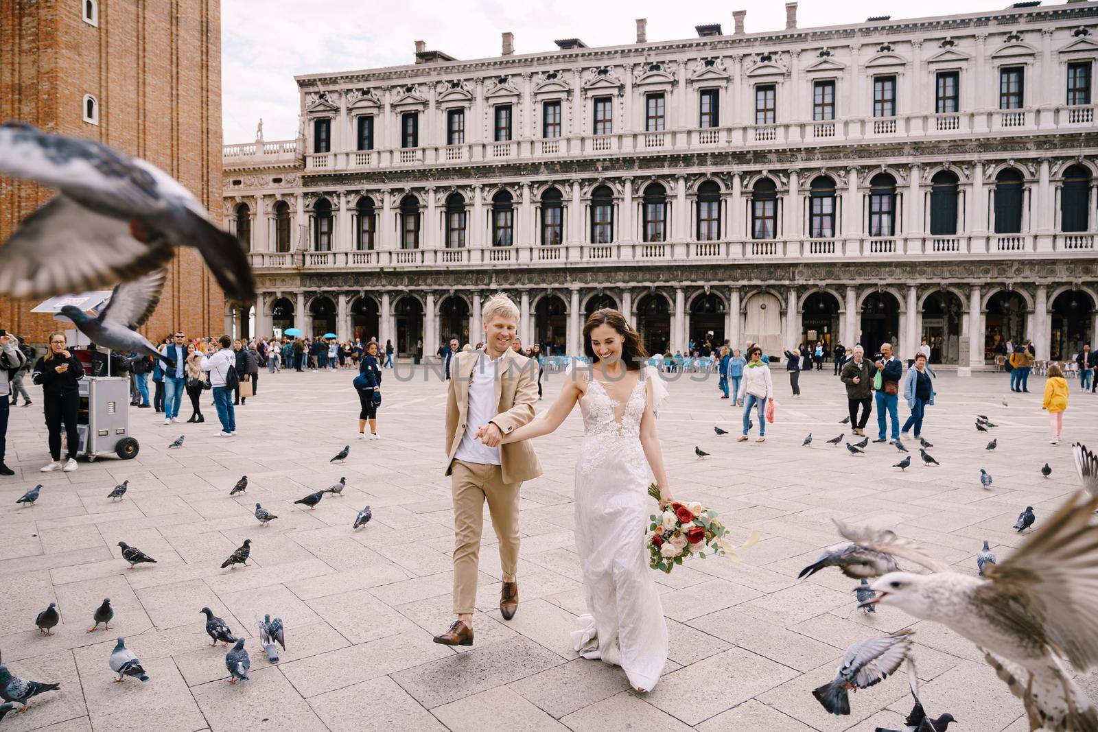 Venice, Italy - 04 october 2019: Venice Wedding, Italy. The bride and groom are running through a flock of flying pigeons in Piazza San Marco, amid the National Archaeological Museum Venice by Nadtochiy