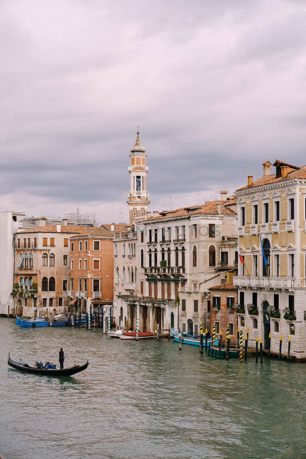 A gondola with tourists is sailing along Grand Canal amid the facades of Venetian houses standing on the water in Venice, Italy.