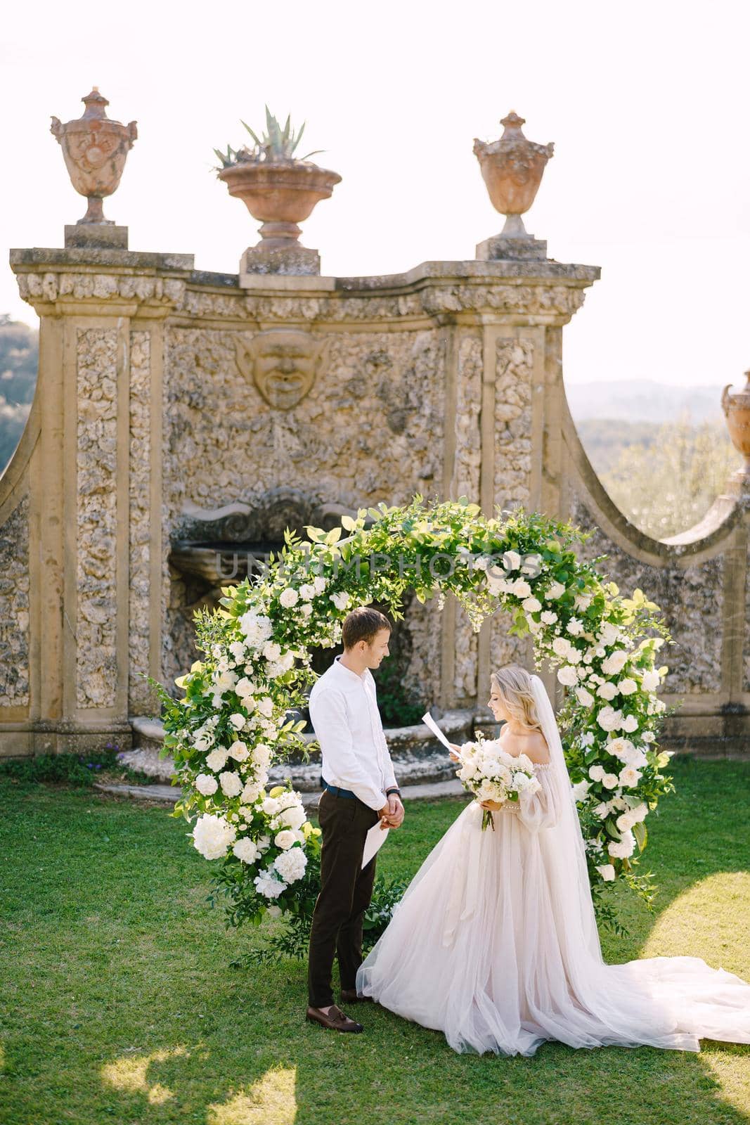 The bride reads wedding vows. Wedding at an old winery villa in Tuscany, Italy. Round wedding arch decorated with white flowers and greenery in front of an ancient Italian architecture. by Nadtochiy