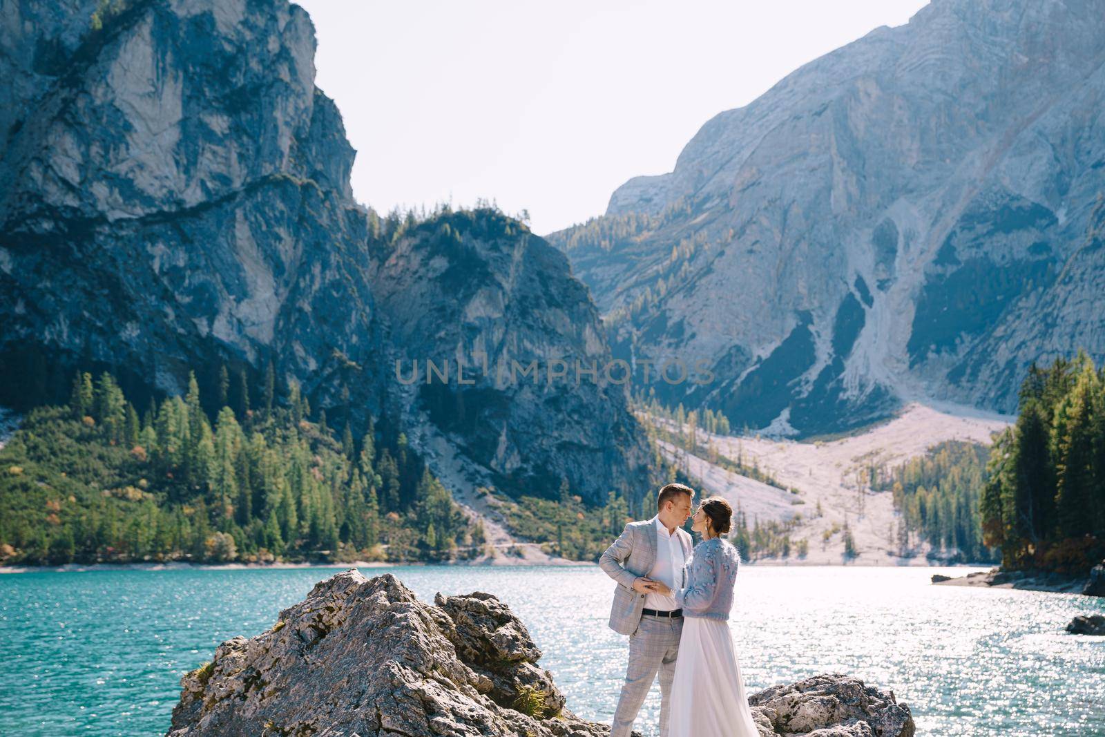 The bride and groom are standing on stones overlooking the Lago di Braies in Italy. Destination wedding in Europe, on Braies lake. Loving newlyweds walk against the backdrop of amazing nature. by Nadtochiy