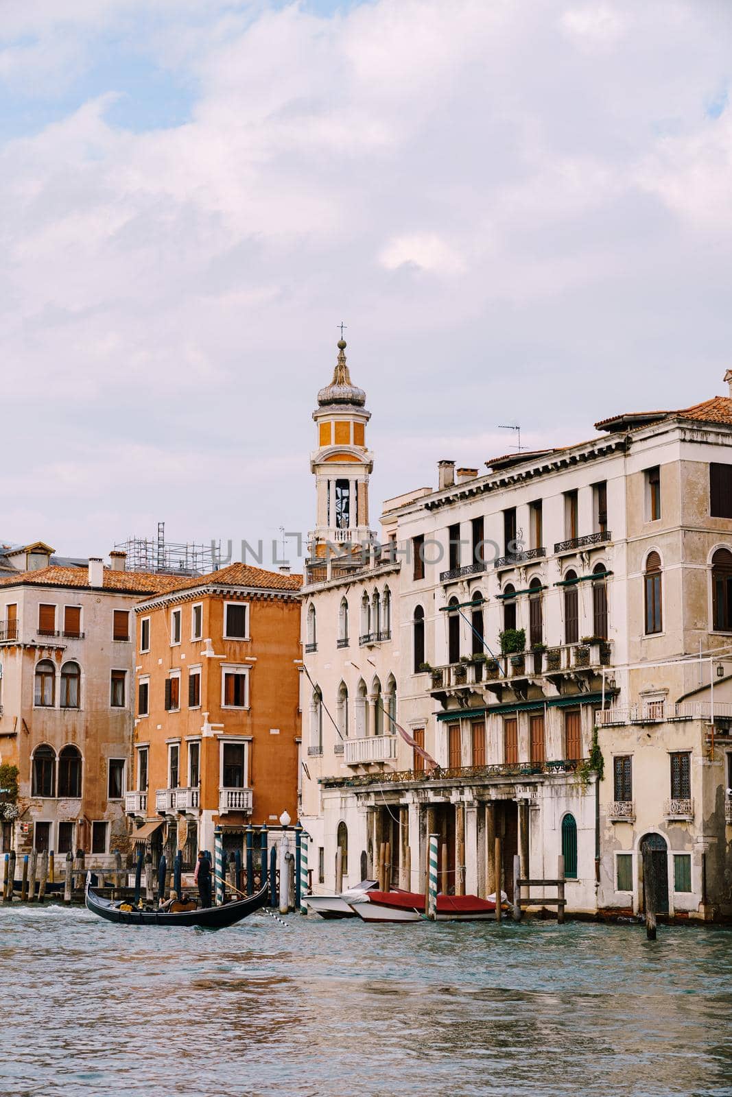 A gondola with tourists is sailing along Grand Canal amid the facades of Venetian houses standing on the water in Venice, Italy.