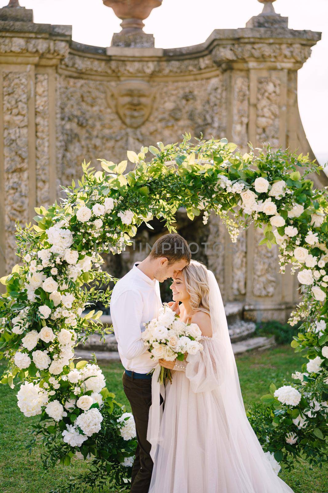 Wedding couple near round wedding arch decorated with white flowers and greenery in front of an ancient Italian architecture. Wedding at an old winery villa in Tuscany, Italy