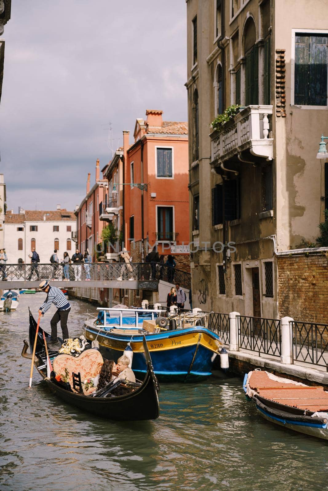 Venice, Italy - 04 october 2019: Italy wedding in Venice. A gondolier rolls a bride and groom in a classic wooden gondola along a narrow Venetian canal. Newlyweds are sitting in a boat and kissing. by Nadtochiy