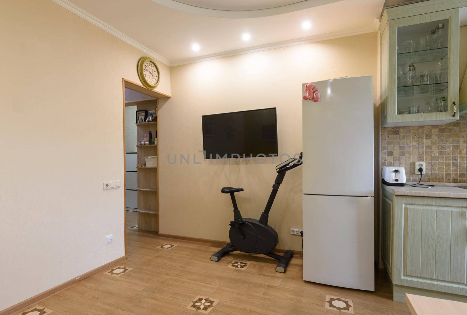 A fragment of the interior of the living room combined with the kitchen, a TV hangs on the wall, an exercise bike and a refrigerator are located nearby