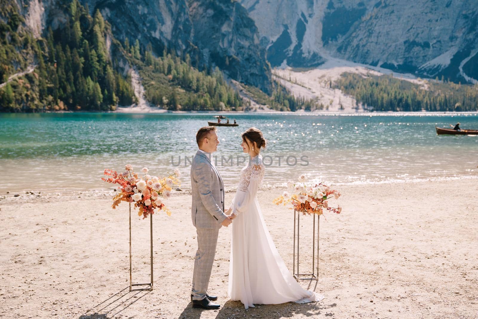 The groom dons a bride's ring, in place for the ceremony, with an arch of autumn floral columns, against the backdrop of Lago di Braies in Italy. Destination wedding in Europe, at Braies lake.