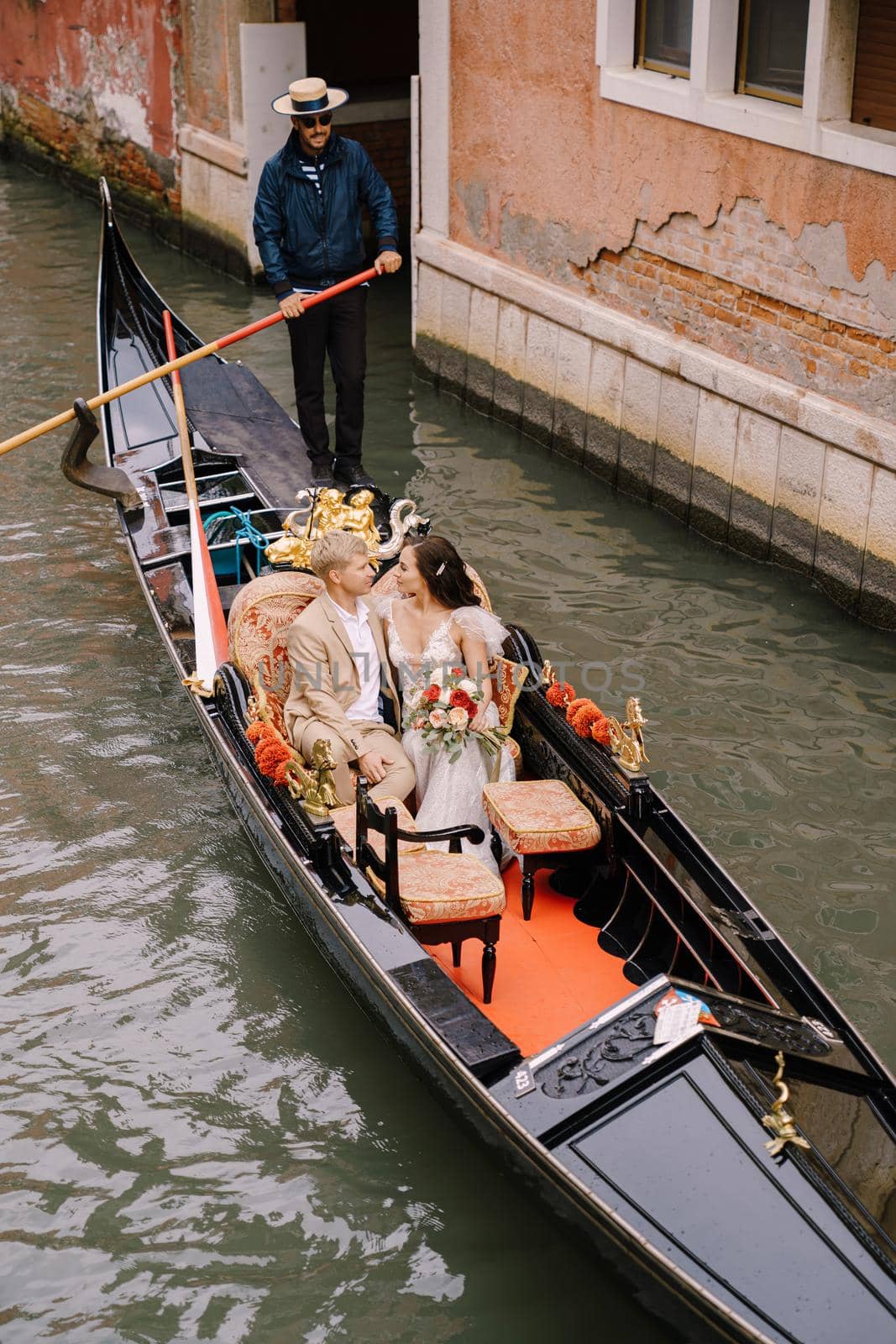 The gondolier rides the bride and groom in a classic wooden gondola along a narrow Venetian canal. Newlyweds sit in a boat and want to kiss.