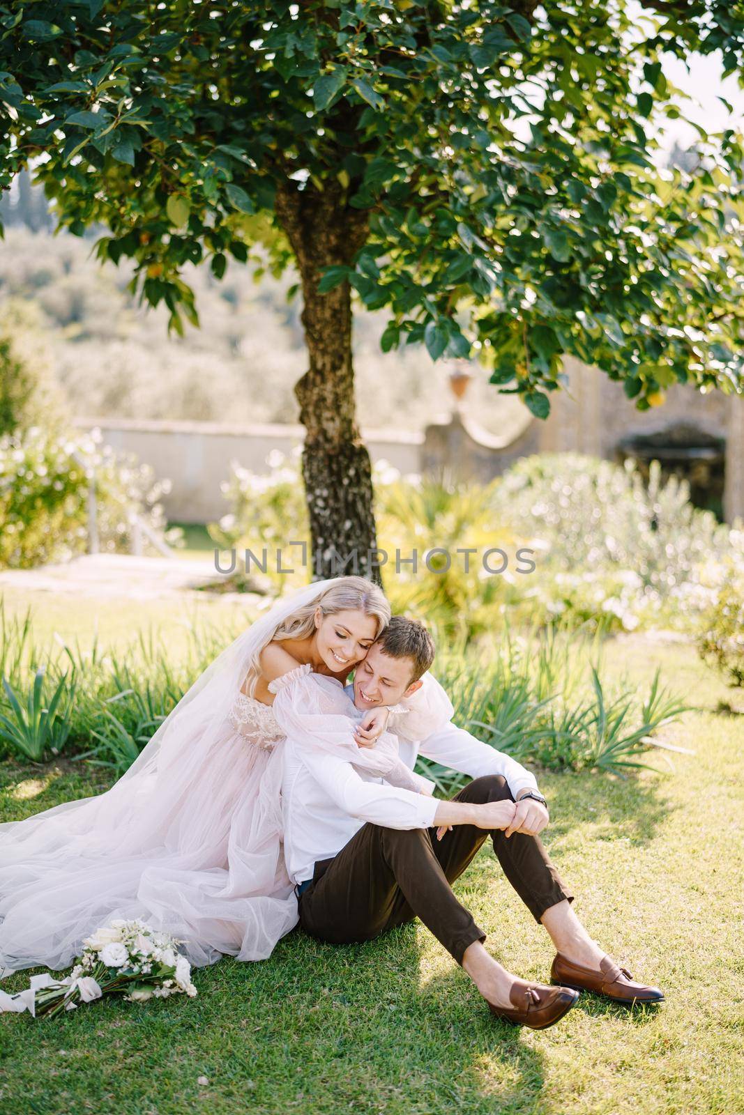 Wedding in Florence, Italy, in an old villa-winery. A wedding couple is sitting on the grass in the garden under a tree, the bride is hugging the groom. by Nadtochiy