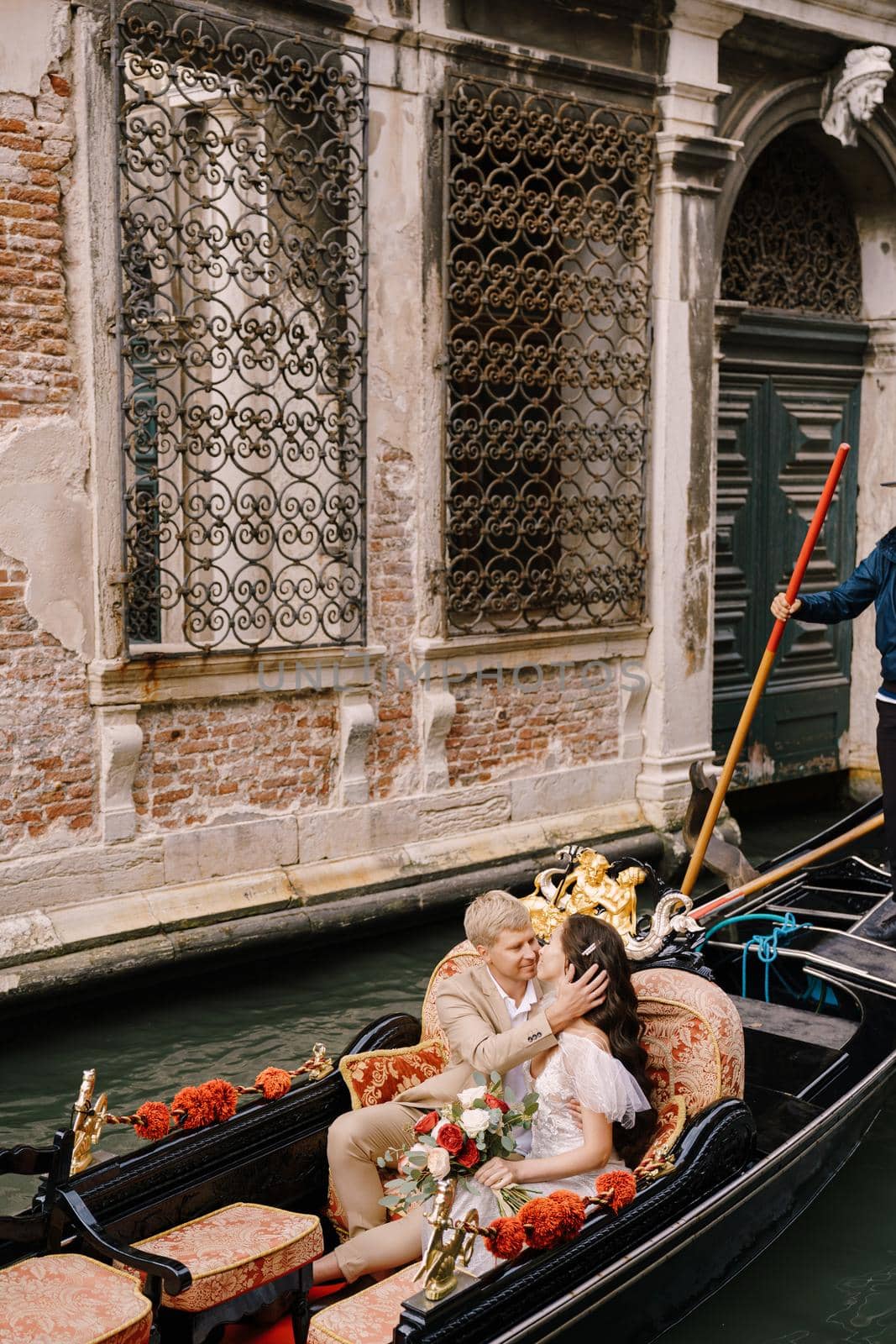 The gondolier rides the bride and groom in a classic wooden gondola along a narrow Venetian canal. Newlyweds sit in a boat spout to the spout, swim against the background of an old forged lattice.