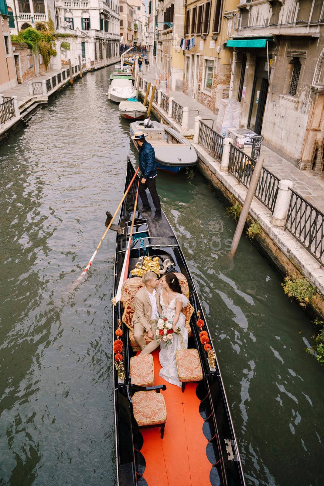 The gondolier rides the bride and groom in a classic wooden gondola along a narrow Venetian canal. Newlyweds sit in a boat against the background of ancient buildings.