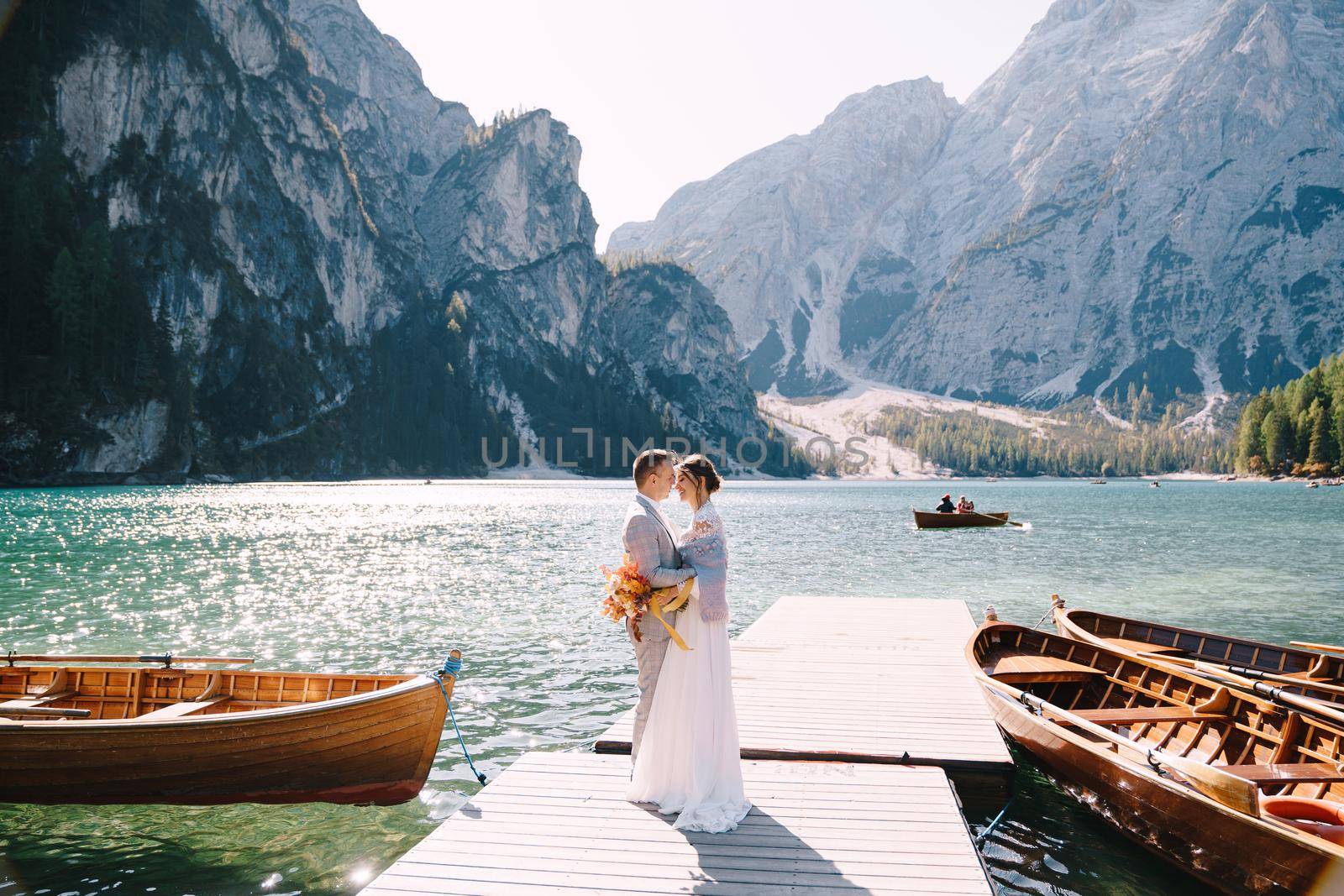 The bride and groom walk on a wooden boat dock at Lago di Braies in Italy. Wedding in Europe, at Braies lake. Newlyweds walk, kiss, cuddle against the backdrop of rocky mountains.