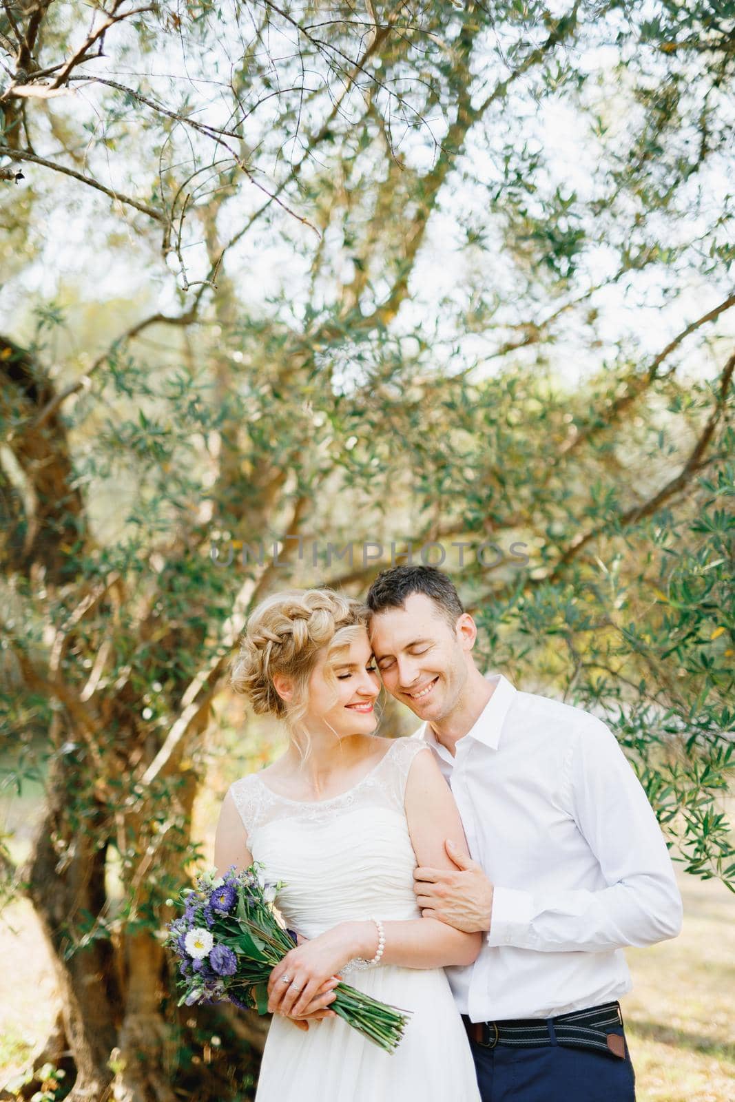 The groom gently hugs the bride near the green trees in the olive grove, the bride holds a bouquet of blue flowers by Nadtochiy