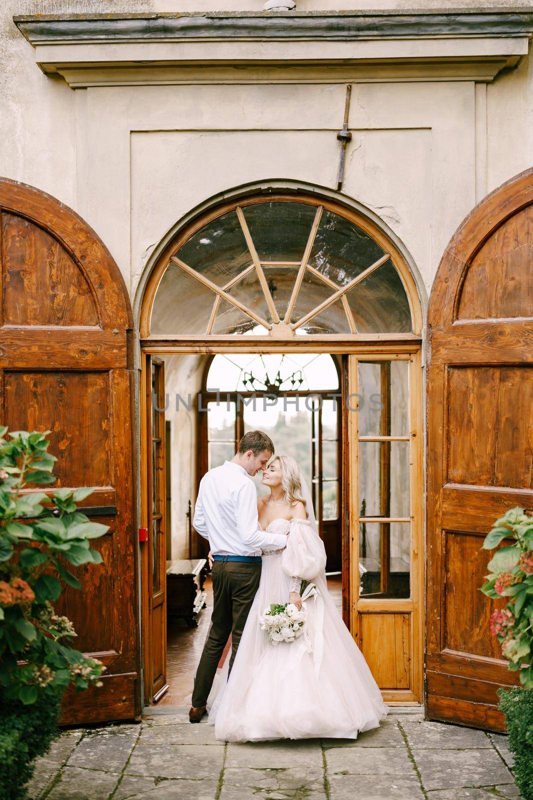 Wedding at an old winery villa in Tuscany, Italy. A wedding couple stands near the old wooden doors at the villa-winery.