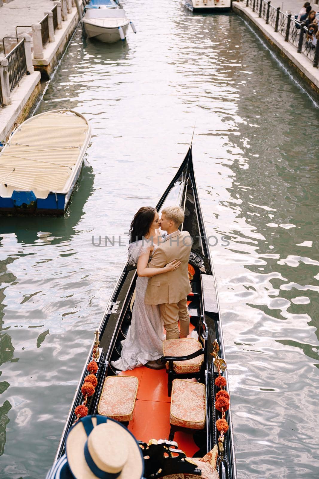 The gondolier rides the bride and groom in a classic wooden gondola along a narrow Venetian canal. The newlyweds stand in the boat, the view from behind.