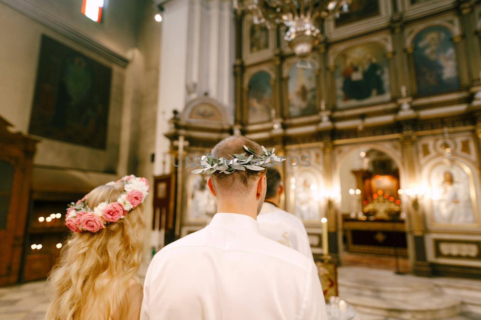 Budva, Montenegro - 21 august 2020: The priest, the groom and the bride in wreaths stand at the altar of the Church of St. Nicholas in Kotor during the wedding, back view by Nadtochiy