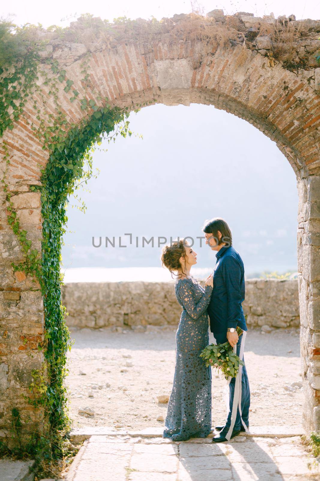 The bride and groom stand side by side in an old arch, the bride put her hands on the groom's chest by Nadtochiy