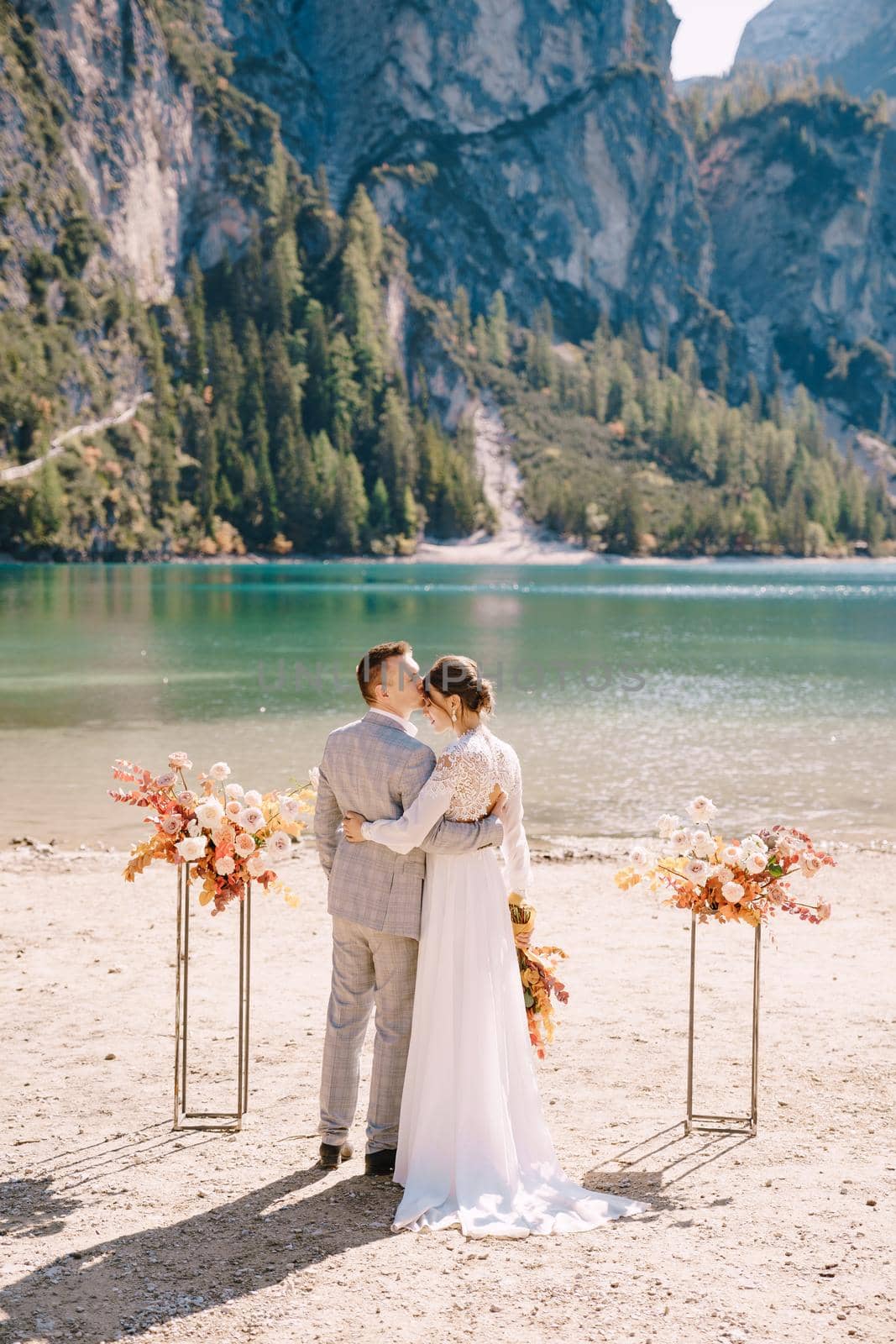 The bride and groom stand still for the ceremony, with an arch of autumn floral columns, against the backdrop of Lago di Braies in Italy. Destination wedding in Europe, at Braies lake.
