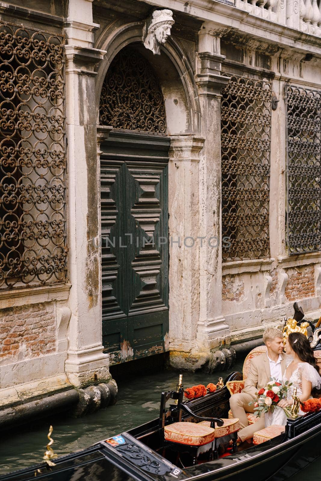 Italy wedding in Venice. Gondolier rolls bride and groom in classic wooden gondola along narrow Venetian canal. Newlyweds are sitting in boat nose to nose, swim against backdrop of old forged lattice by Nadtochiy