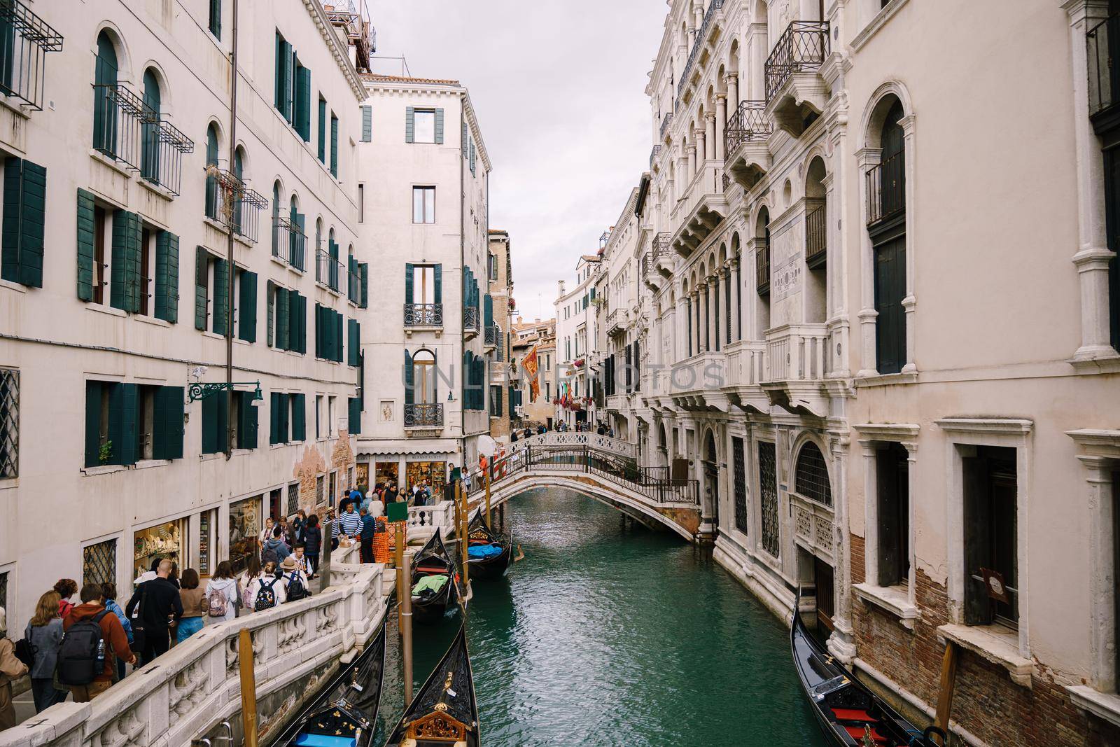 A crowd of tourists walks the streets of Venice, in Italy, near narrow canal between buildings. Small arched bridges over sea water