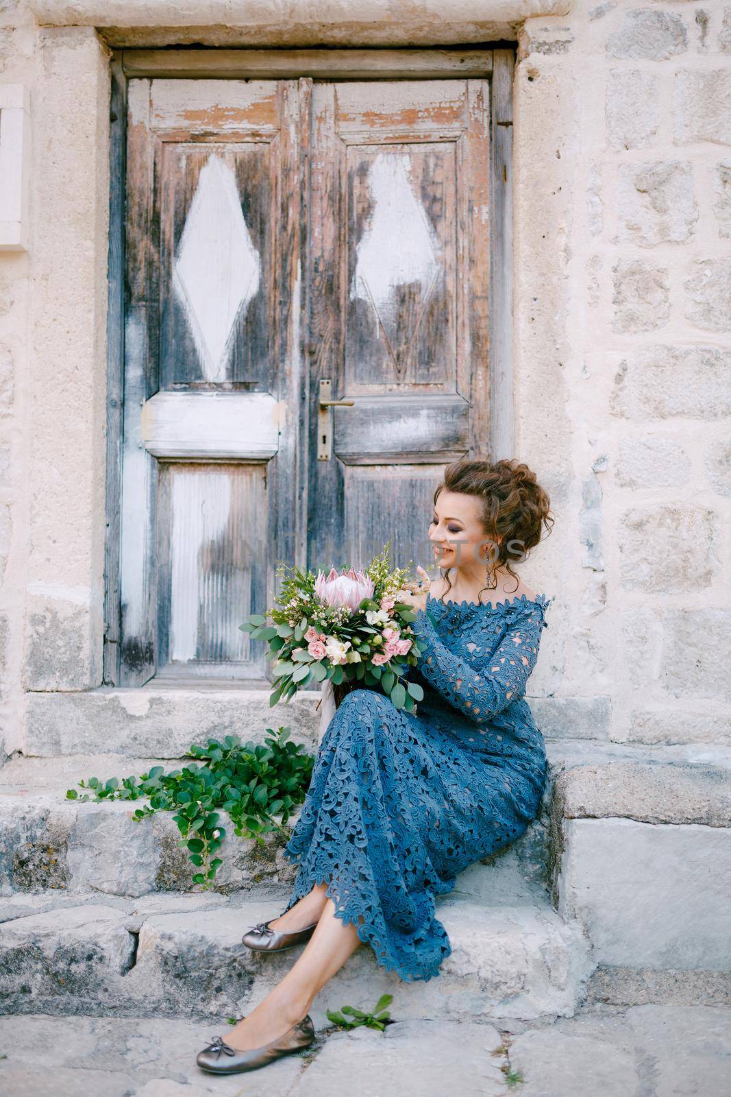 A bride in a stylish blue dress with a bouquet in her hands sits on the steps near an old wooden door . High quality photo
