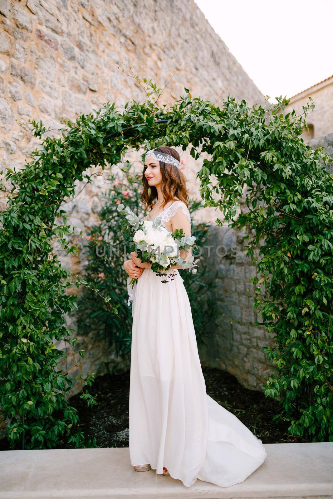 A bride stands at a graceful arch entwined with wild grapes in the old town of Budva and holds a bouquet in her hands . High quality photo