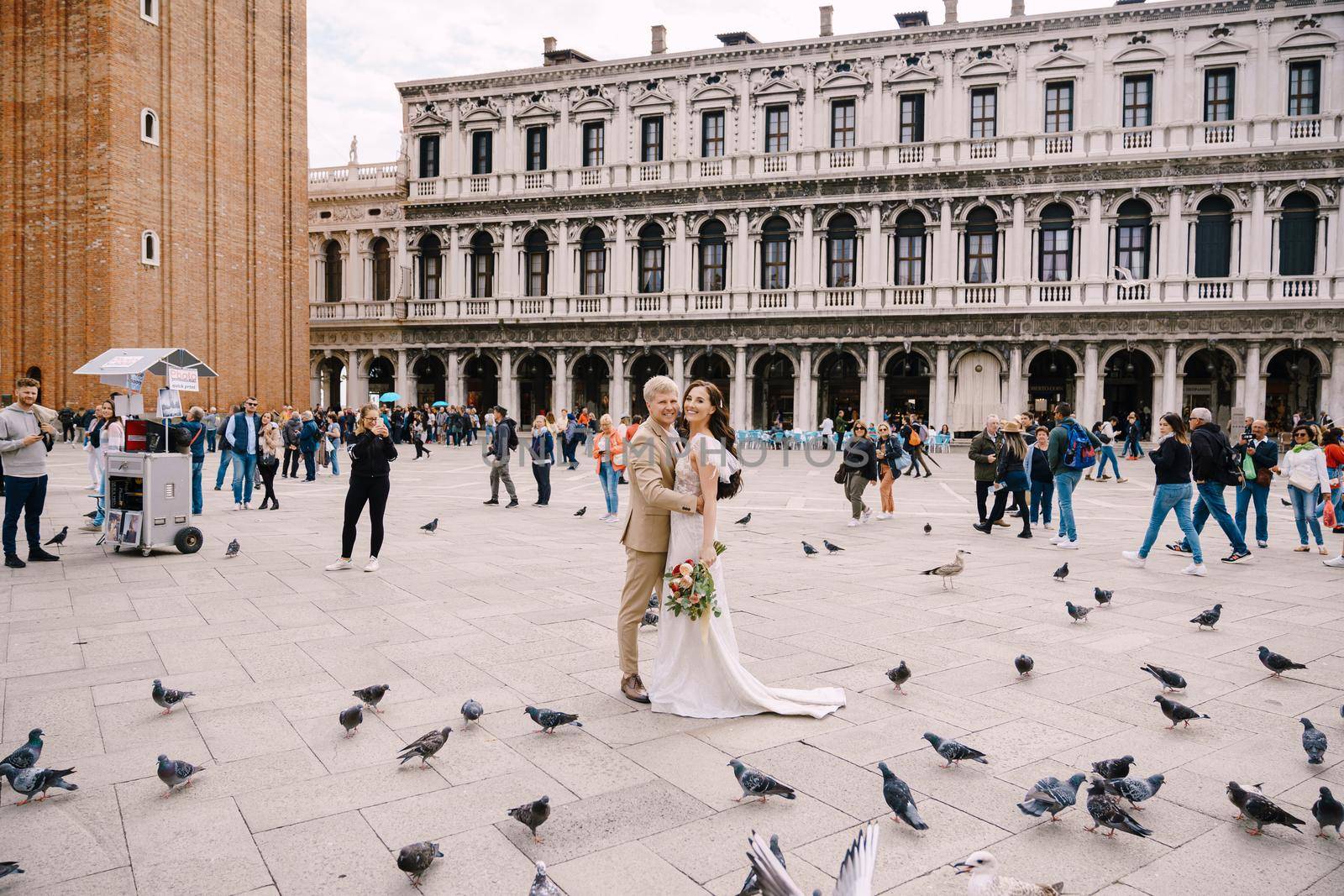 Wedding in Venice, Italy. Bride and groom are looking at camera among the pigeons in Piazza San Marco, against backdrop of National Archaeological Museum Venice, surrounded by a crowd of tourists.