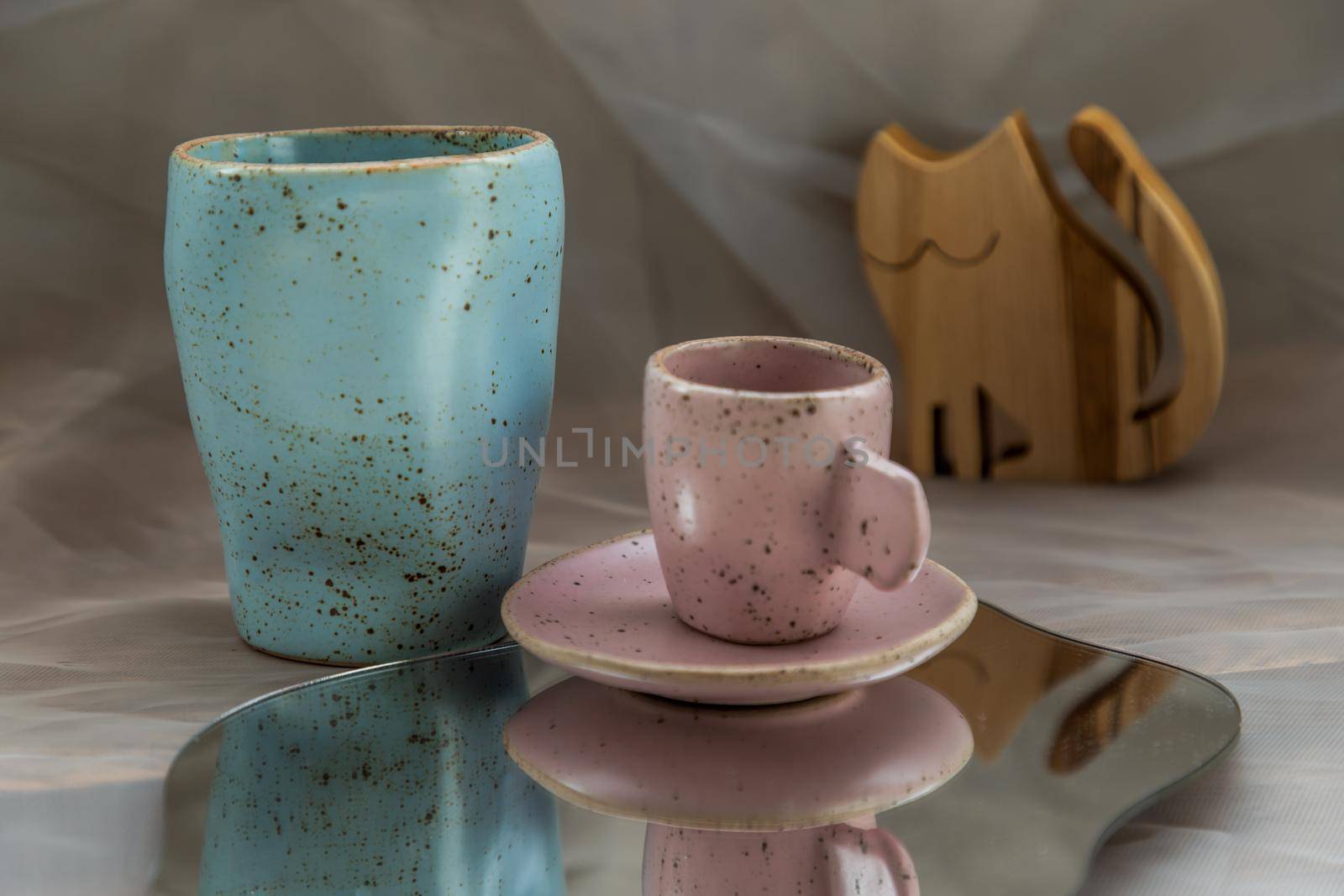 Blue and Pink handmade ceramic mugs on mirror with wooden doll in cat shape on fabric textured table cloth. Home decor, Copy space, Selective focus.